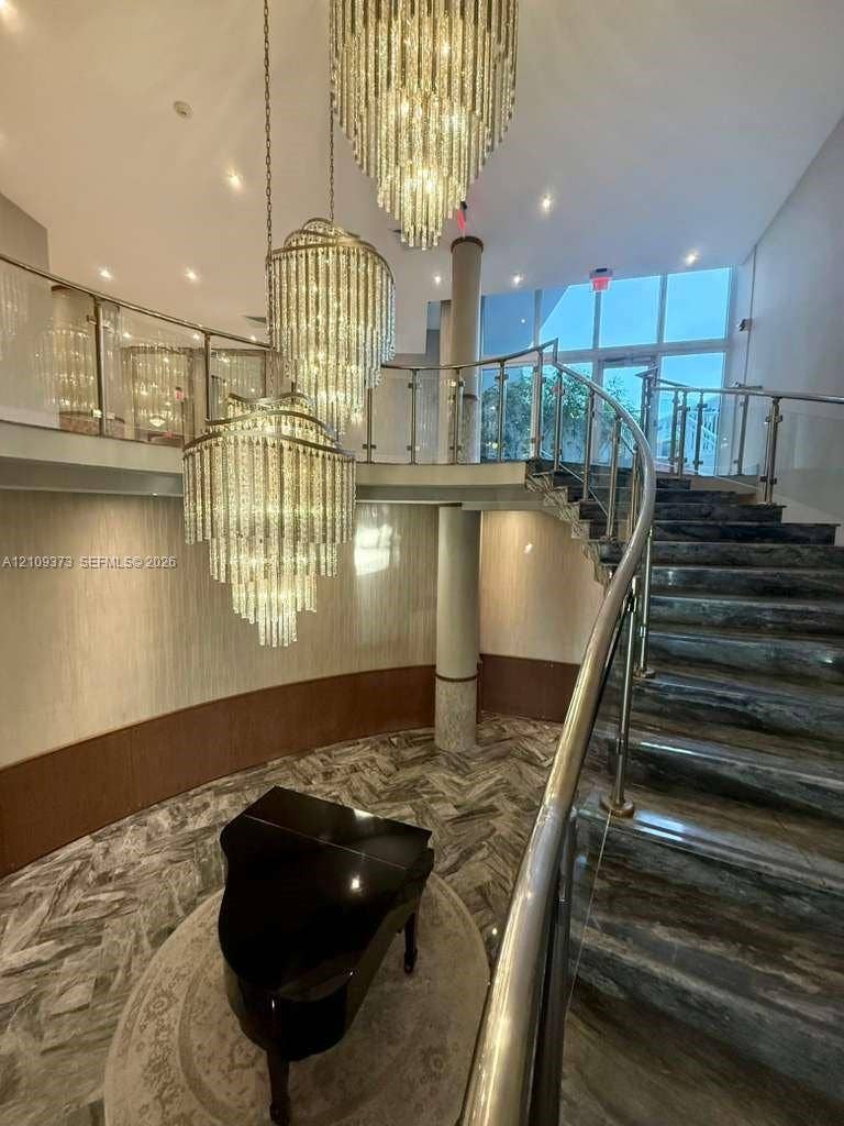 Grand staircase featuring stone-finish steps, a curved glass railing with metal accents, tiered crystal chandeliers, and patterned stone flooring