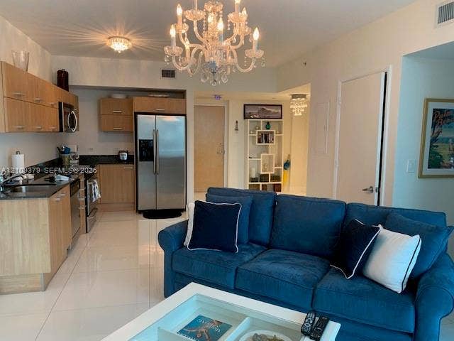 Open-concept interior featuring wood-finish cabinetry, stainless steel appliances, polished stone countertops, ceramic tile flooring, and decorative crystal chandelier lighting