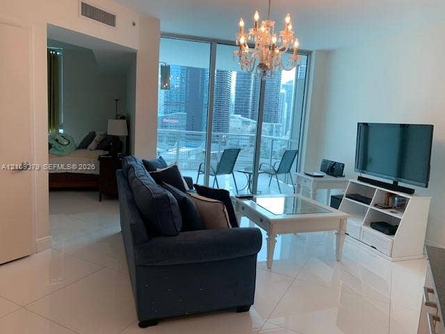 Living space featuring polished tile flooring, floor-to-ceiling glass paneling, a multi-tiered crystal chandelier, and a balcony with metal railing