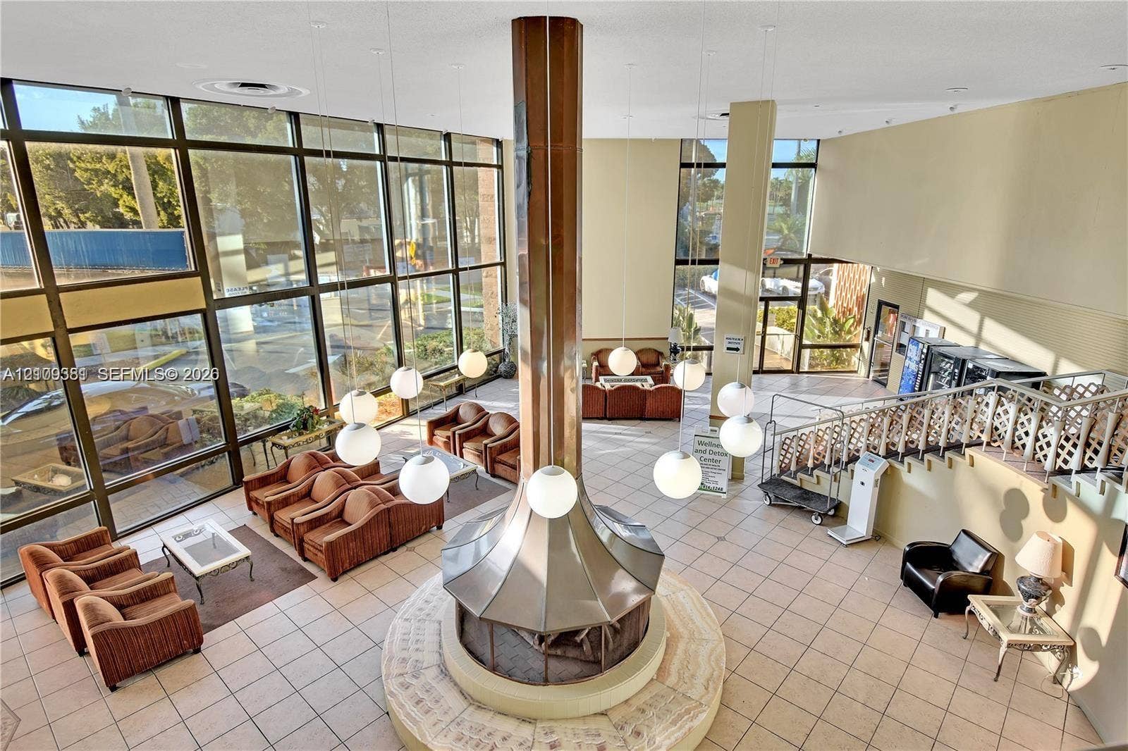 26 Diplomat Parkway, #2351, Hallandale Beach, FL 33009 Floor-to-ceiling multi-pane windows, central cylindrical fireplace with metallic finish, globe pendant lighting, ceramic tile flooring, and decorative metal stair railing