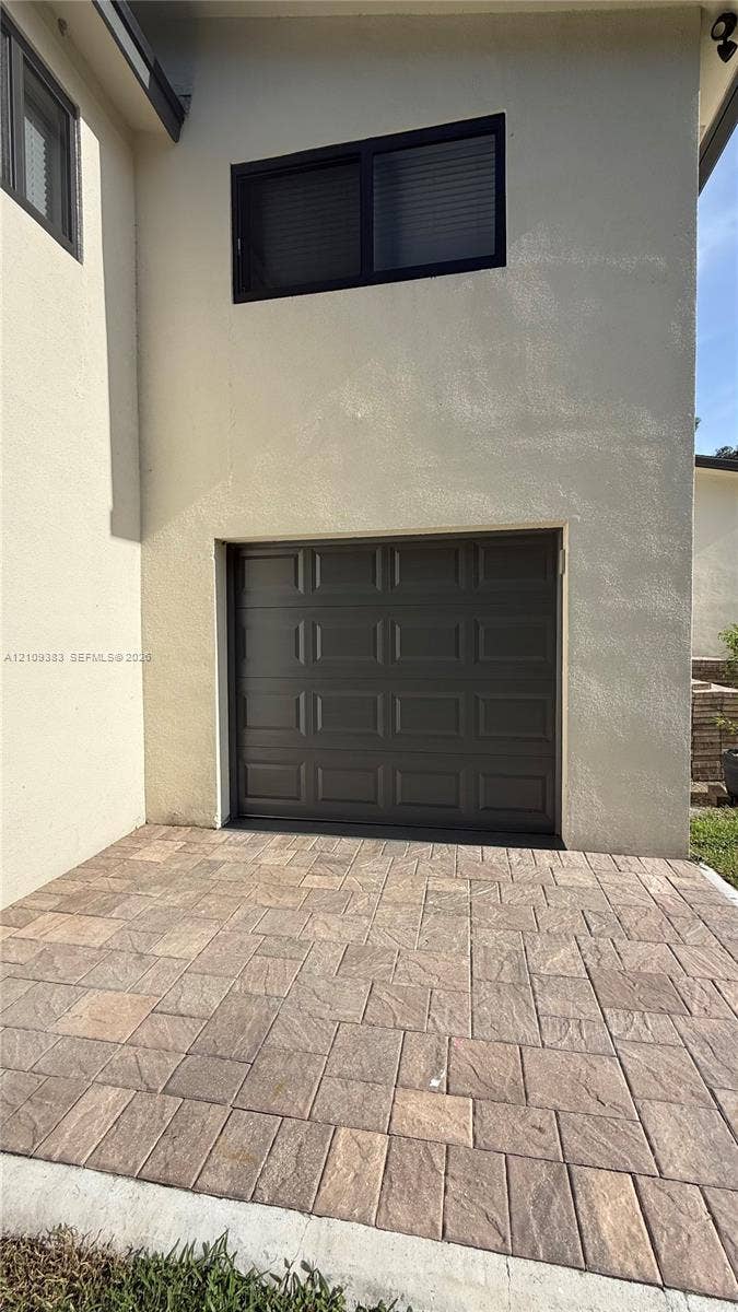 10751 SW 48th Street, Davie, Davie, FL 33328 Stucco exterior featuring a single-car garage with a paneled door, a rectangular window with a dark frame, and a patterned paver driveway