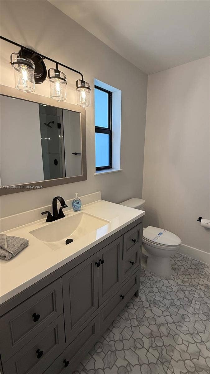 10751 SW 48th Street, Davie, Davie, FL 33328 Gray vanity cabinet with a light-toned countertop, undermount sink, matte black faucet, and patterned tile flooring, complemented by a three-light fixture and framed mirror