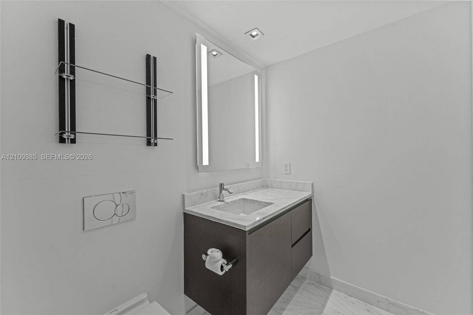 Wall-mounted dark wood-finish vanity with stone countertop, integrated sink, LED-lit mirror, built-in glass shelving, and wall-mounted dual-flush actuator