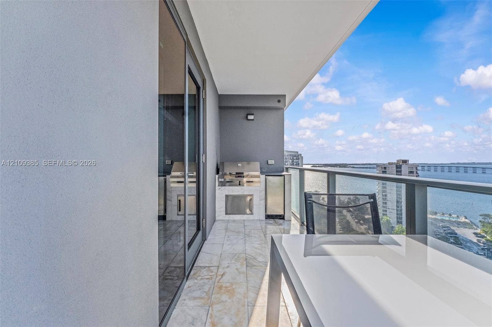 Covered balcony featuring stone-finish flooring, built-in stainless steel outdoor kitchen with stone-veneer base, and glass-paneled railing