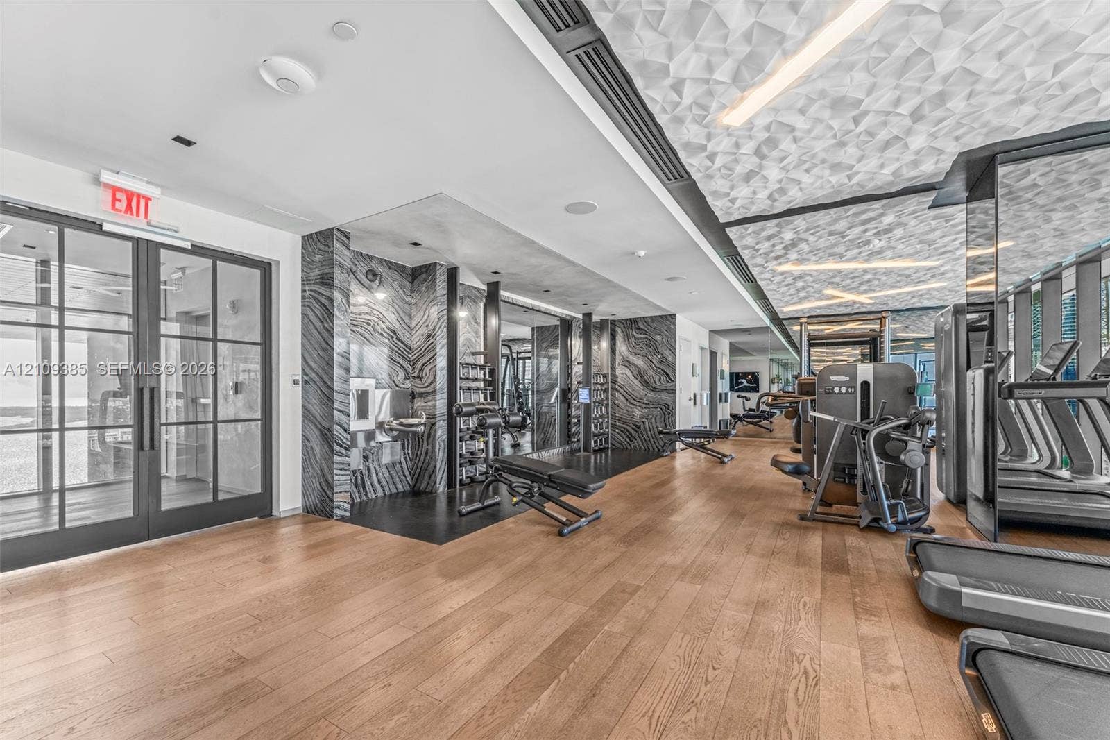 Fitness area featuring wood-finish flooring, floor-to-ceiling black-and-white stone wall cladding, a textured geometric ceiling, and double-paned glass-panel doors