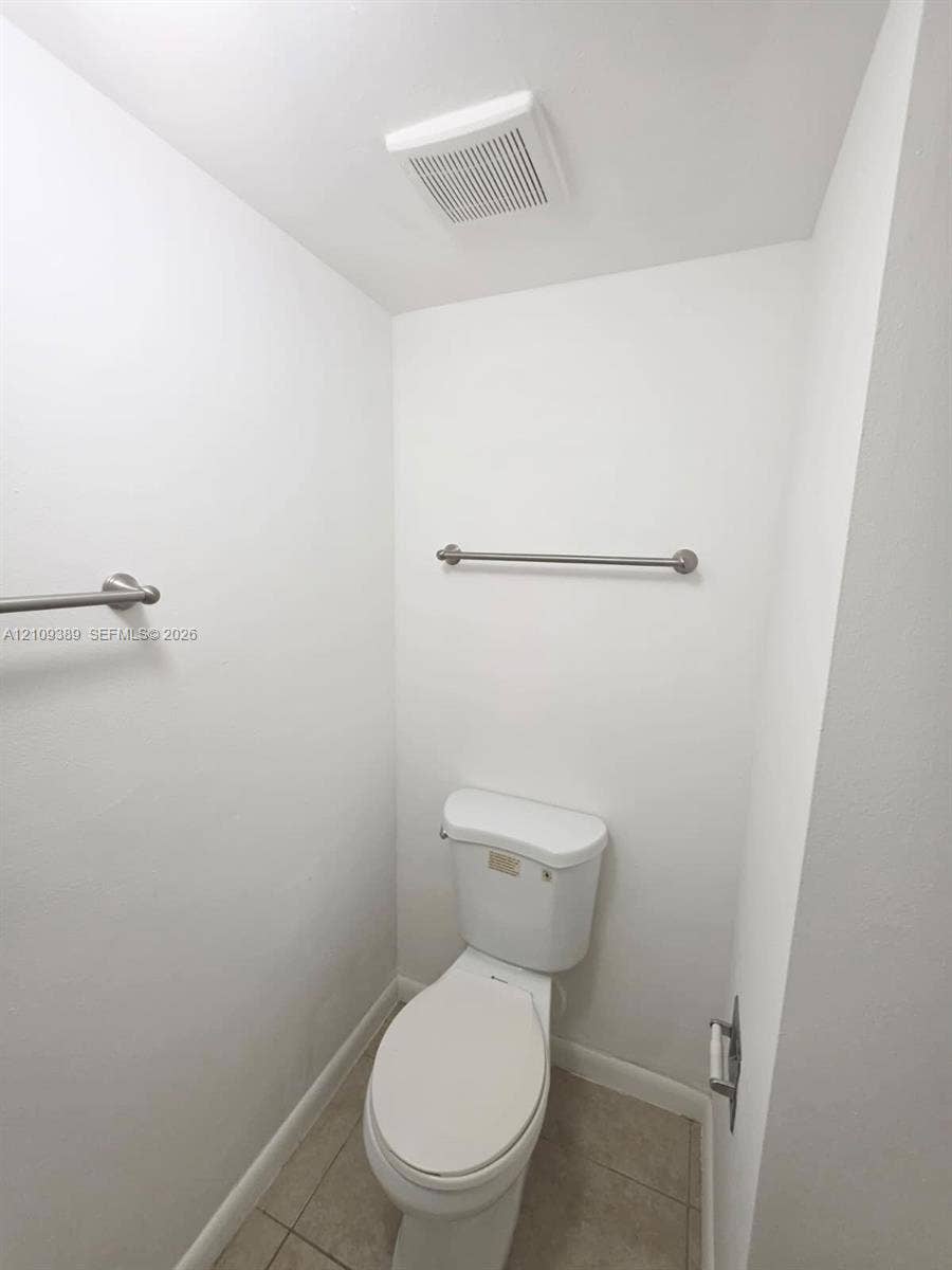 White toilet with a matching elongated seat, tile flooring, and brushed metal hardware including wall-mounted towel bars and a toilet paper holder