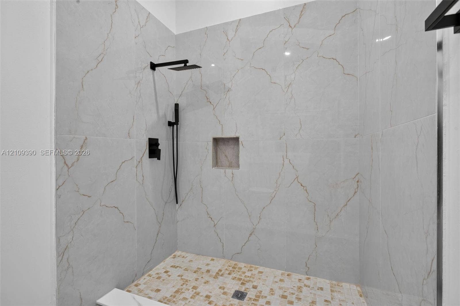 Walk-in shower featuring large-format stone-look wall tiling with veining detail, mosaic tile flooring, built-in recessed wall niche, and matte black rainfall showerhead with hand-held sprayer