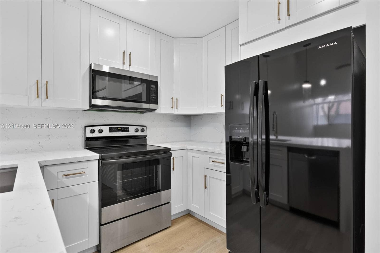 Kitchen featuring white shaker-style cabinetry with gold-tone hardware, white stone-composite countertops, wood-finish flooring, and black-finish stainless steel appliances