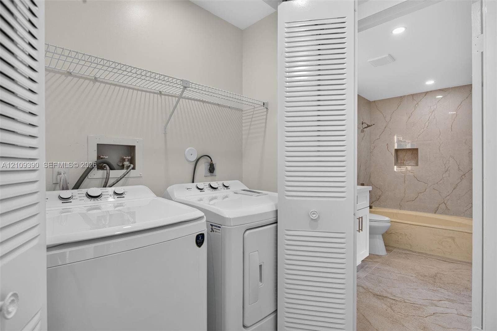 Laundry area featuring wire shelving and accordion-style louvered closet doors, with tile flooring and a tub-shower enclosure finished in a textured stone-look tile