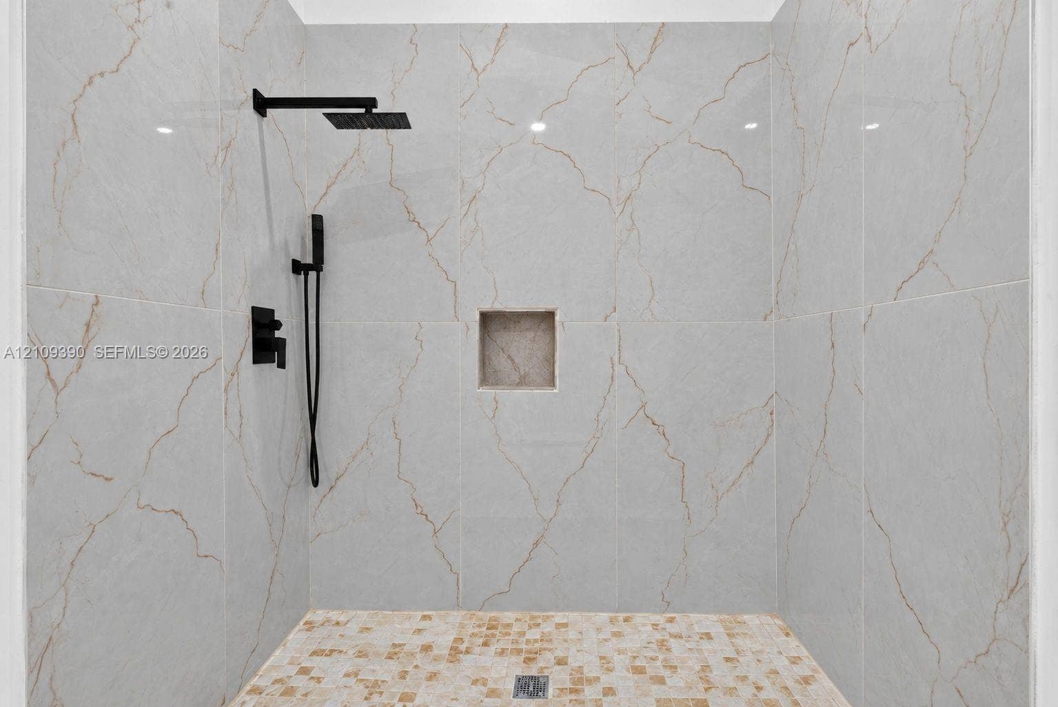 Walk-in shower featuring floor-to-ceiling large-format veined tile, mosaic floor tiling, integrated wall niche, and matte black rainfall showerhead with hand-held sprayer
