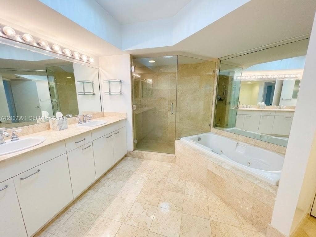 Dual-sink vanity with stone countertop, walk-in glass-enclosed shower, soaking tub with stone tile surround, stone flooring, and mirrored wall surfaces