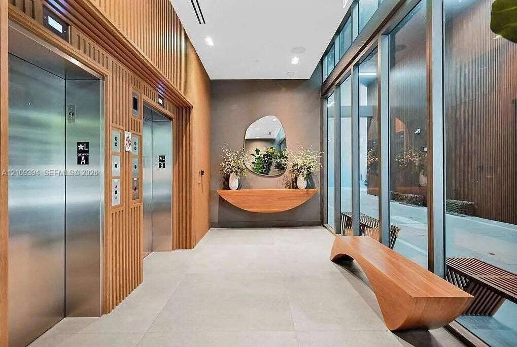 Lobby area featuring stainless steel elevator doors, floor-to-ceiling glass paneling, wood-slat wall paneling, large-format tile flooring, and a built-in wood console