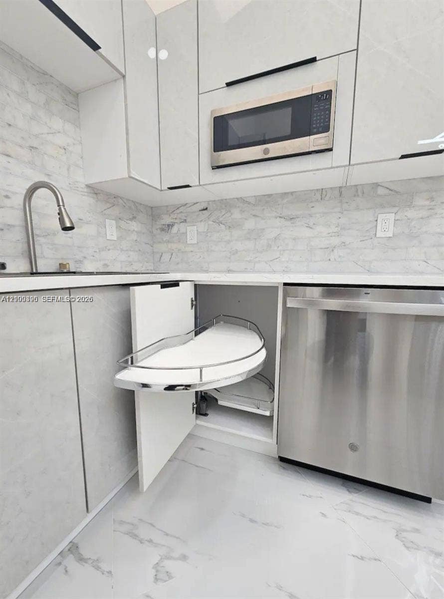Modern kitchen featuring white flat-panel cabinetry, pull-out corner shelving, stainless steel appliances, stone-patterned backsplash, and matching stone-patterned flooring