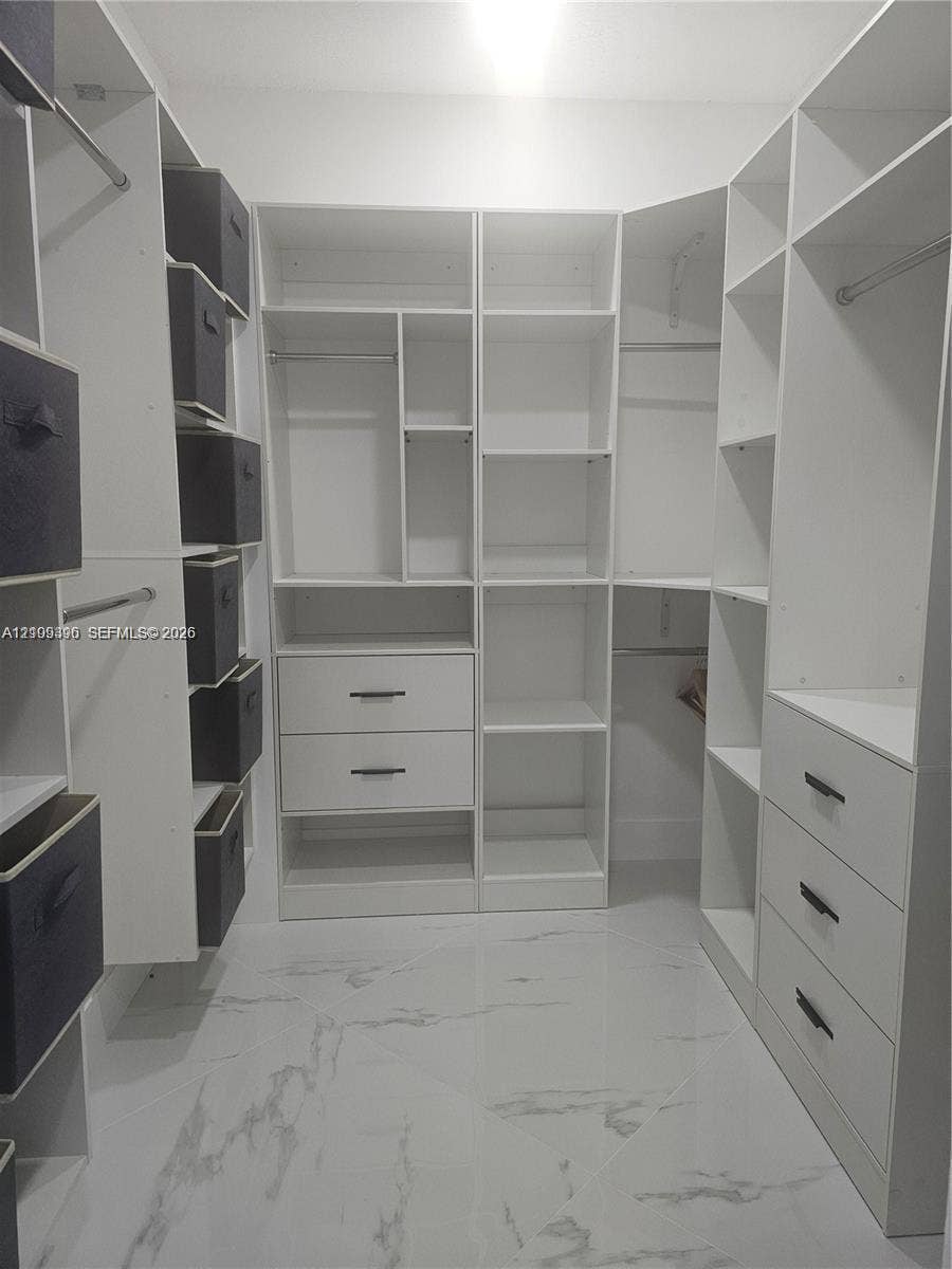Walk-in closet featuring custom white cabinetry, built-in shelving, integrated dresser drawers with matte black pulls, and large-format polished flooring with a marble-finish aesthetic