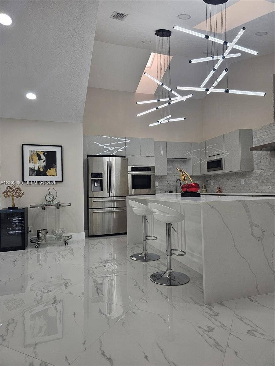 Modern kitchen featuring a waterfall island with stone-finish countertops, high-gloss cabinetry, stainless steel appliances, large-format tile flooring, and geometric multi-pendant lighting