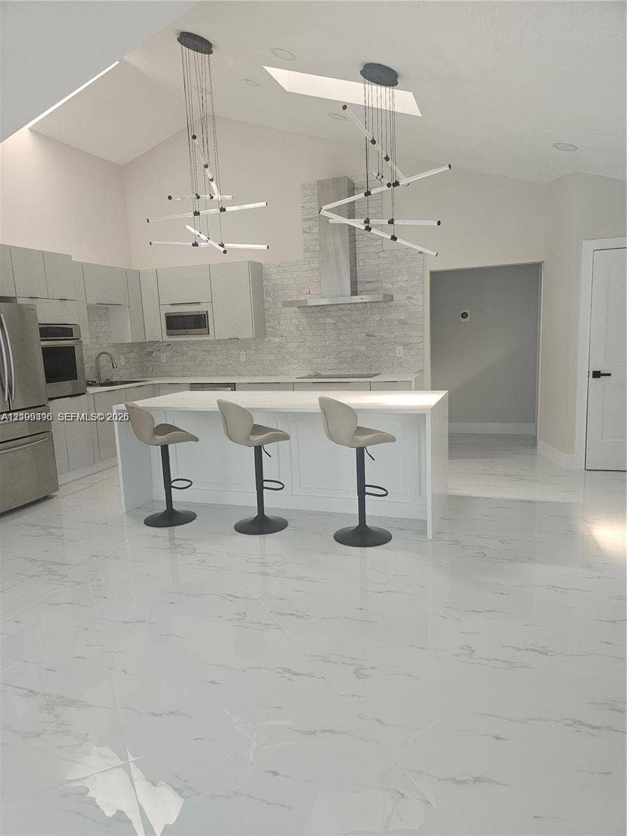 Kitchen featuring vaulted ceilings, skylights, white cabinetry, a center island with solid surfacing, and a stone-tiled backsplash with a stainless steel range hood