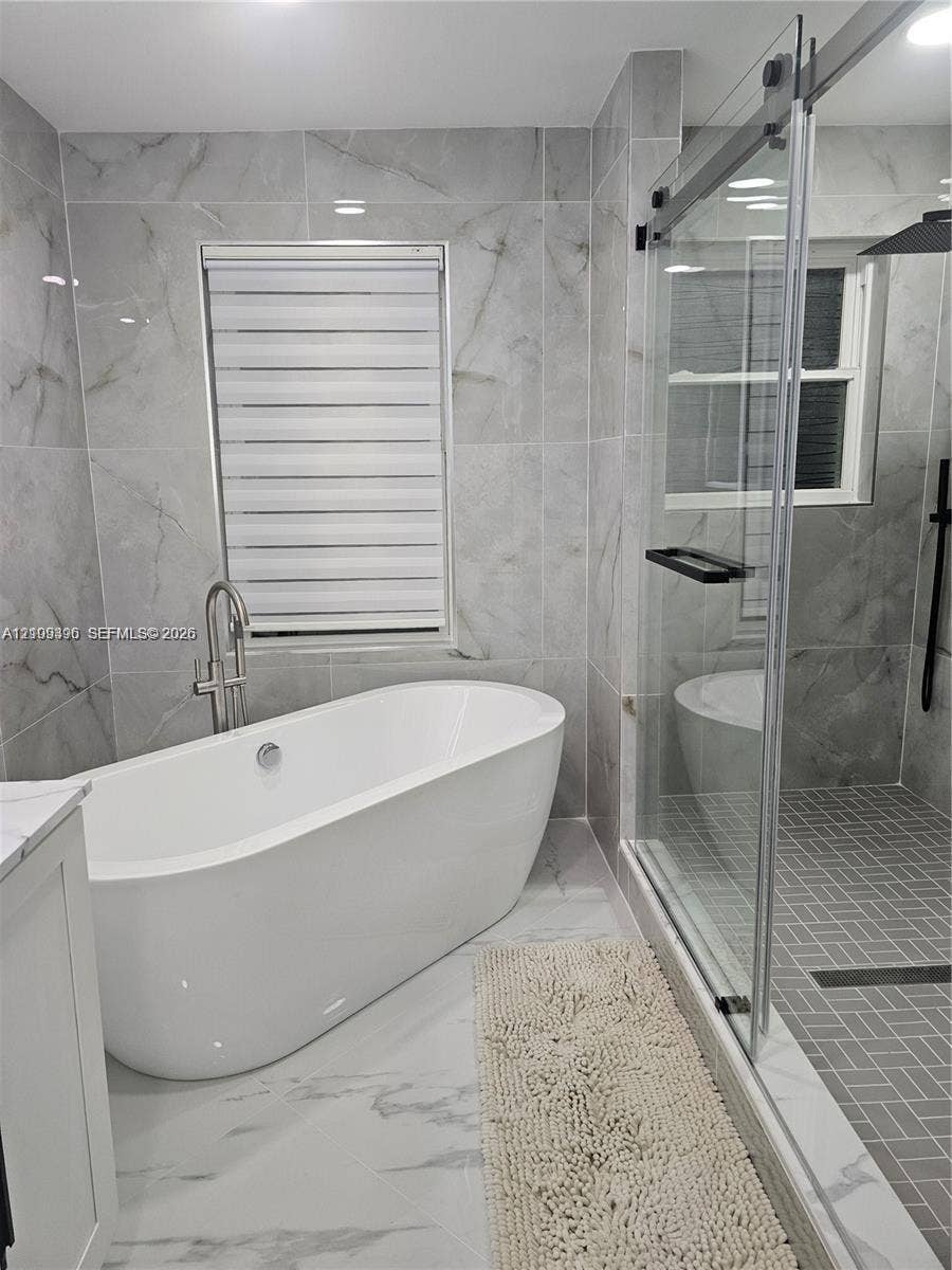 Freestanding soaking tub with a floor-mounted faucet, walk-in shower with a sliding glass door and gray herringbone tile floor, and full-height stone-finish wall tile