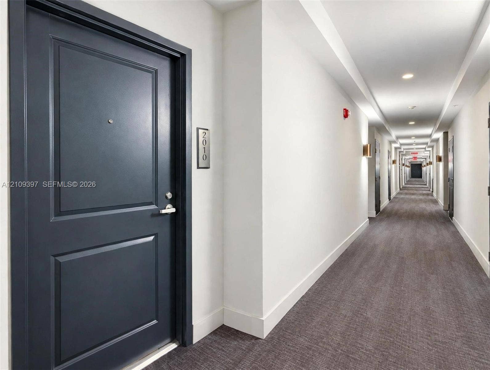 Dark charcoal entry door featuring a brushed metal peephole, metallic door handle, and matching unit number placard, complemented by wall-mounted sconce lighting and neutral-toned carpeting