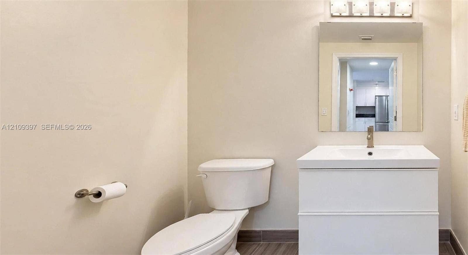 White vanity with integrated rectangular sink and polished chrome faucet, rectangular wall-mounted mirror, four-light vanity fixture, and neutral-tone tile flooring