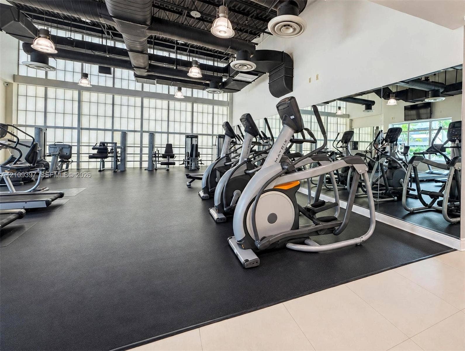 Industrial-style fitness center featuring high ceilings with exposed ductwork, large paneled windows, rubberized flooring, and full-length wall mirrors