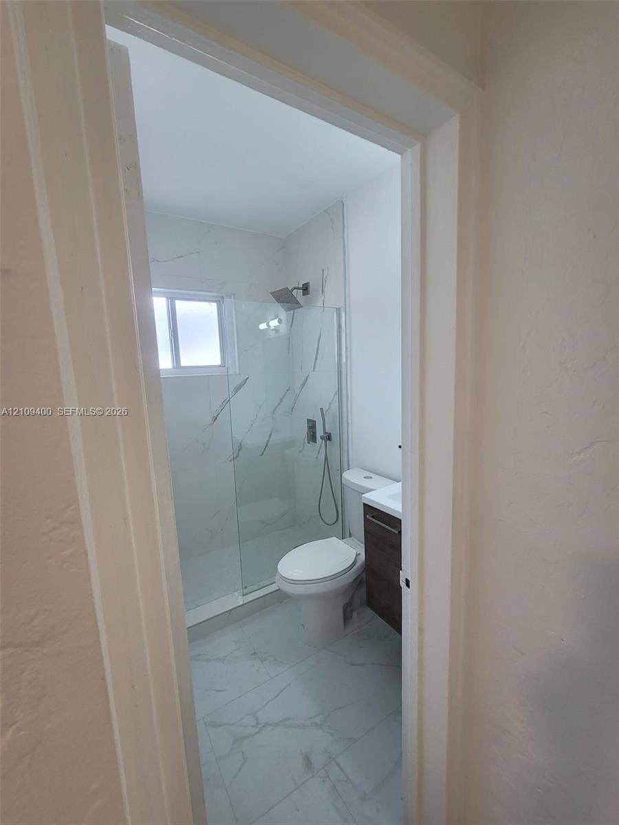 Walk-in shower with frameless glass enclosure, floor-to-ceiling stone-finish wall tile, large-format tile flooring, wall-mounted rainfall showerhead, and wood-finish vanity