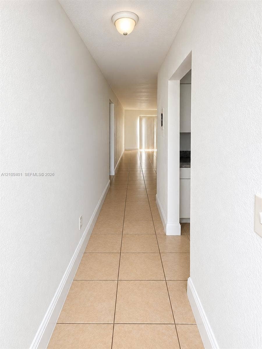 Hallway featuring neutral-toned tile flooring, smooth wall finishes, and a central flush-mount ceiling light fixture