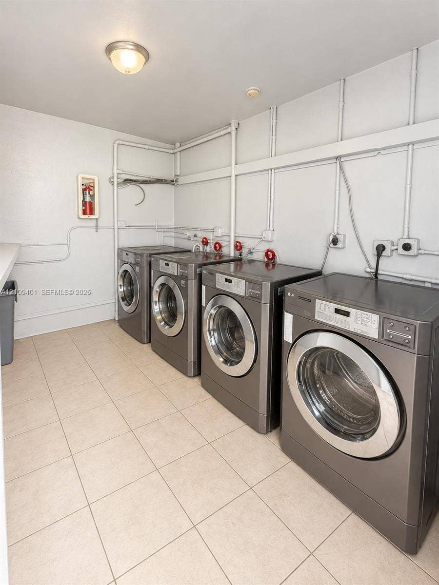 Laundry room featuring four high-capacity front-load washing machines, ceramic tile flooring, and surface-mounted conduit utility lines