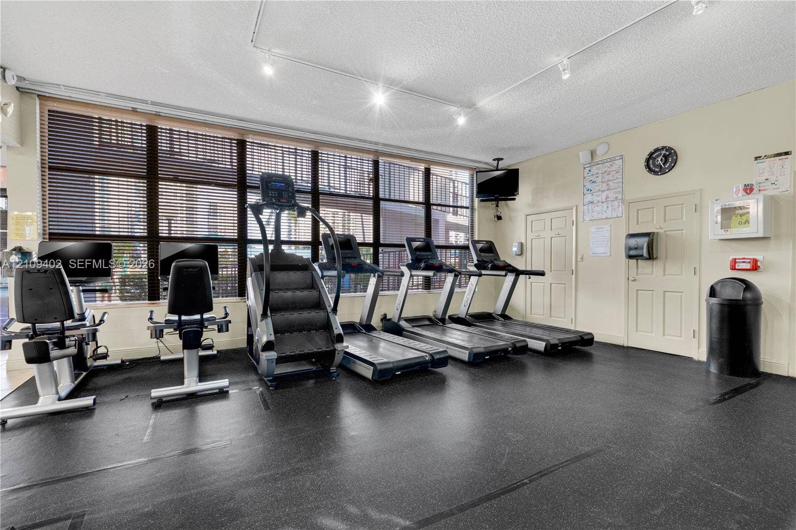 Fitness center featuring wall-to-wall windows with slat blinds, recessed track lighting, and durable rubber flooring