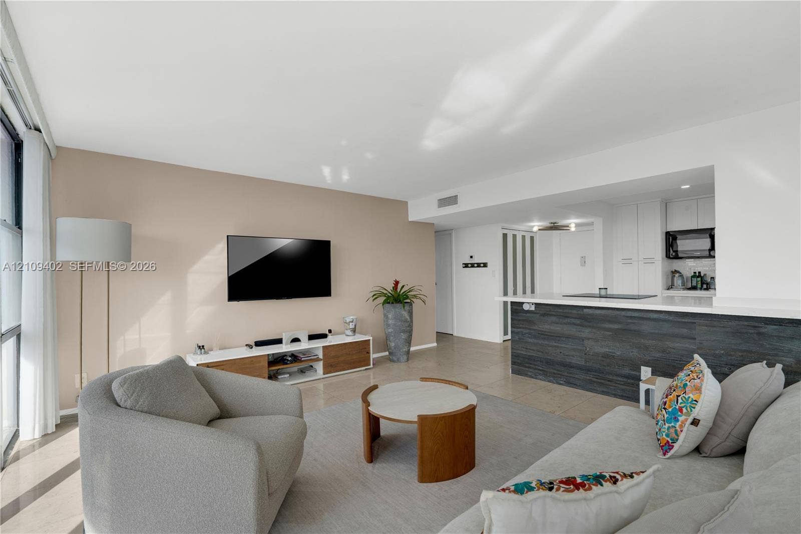 Open-concept interior featuring large-format tile flooring, a kitchen breakfast bar with dark wood-finish paneling, white flat-panel cabinetry, and floor-to-ceiling windows