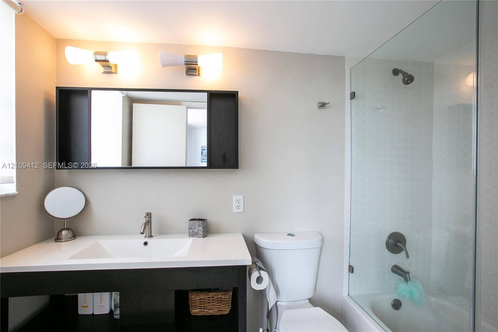 137 Golden Isles Drive, #408, Hallandale Beach, FL 33009 Bathroom featuring a dark-finish vanity with a rectangular sink, white countertop, dual wall-mounted light fixtures, framed medicine cabinet, and a glass-enclosed shower with metallic hardware