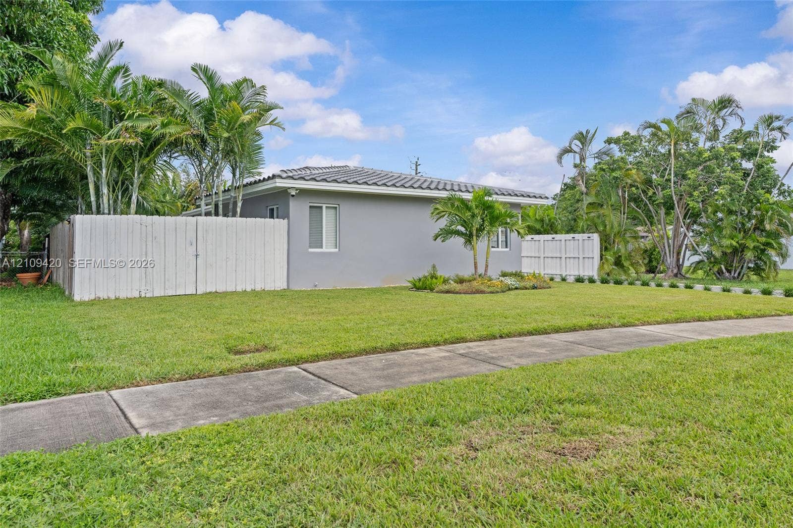 1955 Thomas Street, Hollywood, FL 33020 Single-story exterior with light gray siding, concrete tile roof, white privacy fencing, and established palm landscaping