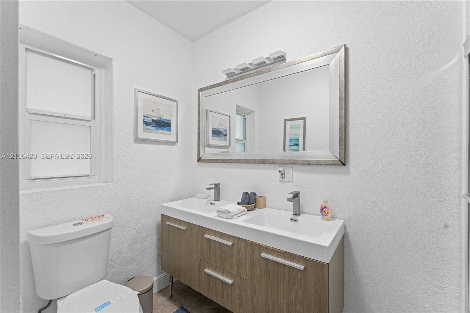 1955 Thomas Street, Hollywood, FL 33020 Dual-sink vanity featuring a wood-finish cabinet, rectangular basin sinks, brushed metal faucets, and a framed wall mirror with integrated lighting