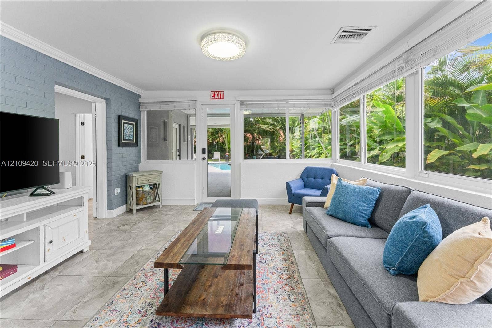 1955 Thomas Street, Hollywood, FL 33020 Sunroom featuring large-format tile flooring, blue-painted brick wall, flush-mount circular lighting, and extensive window enclosures with natural exterior scenery