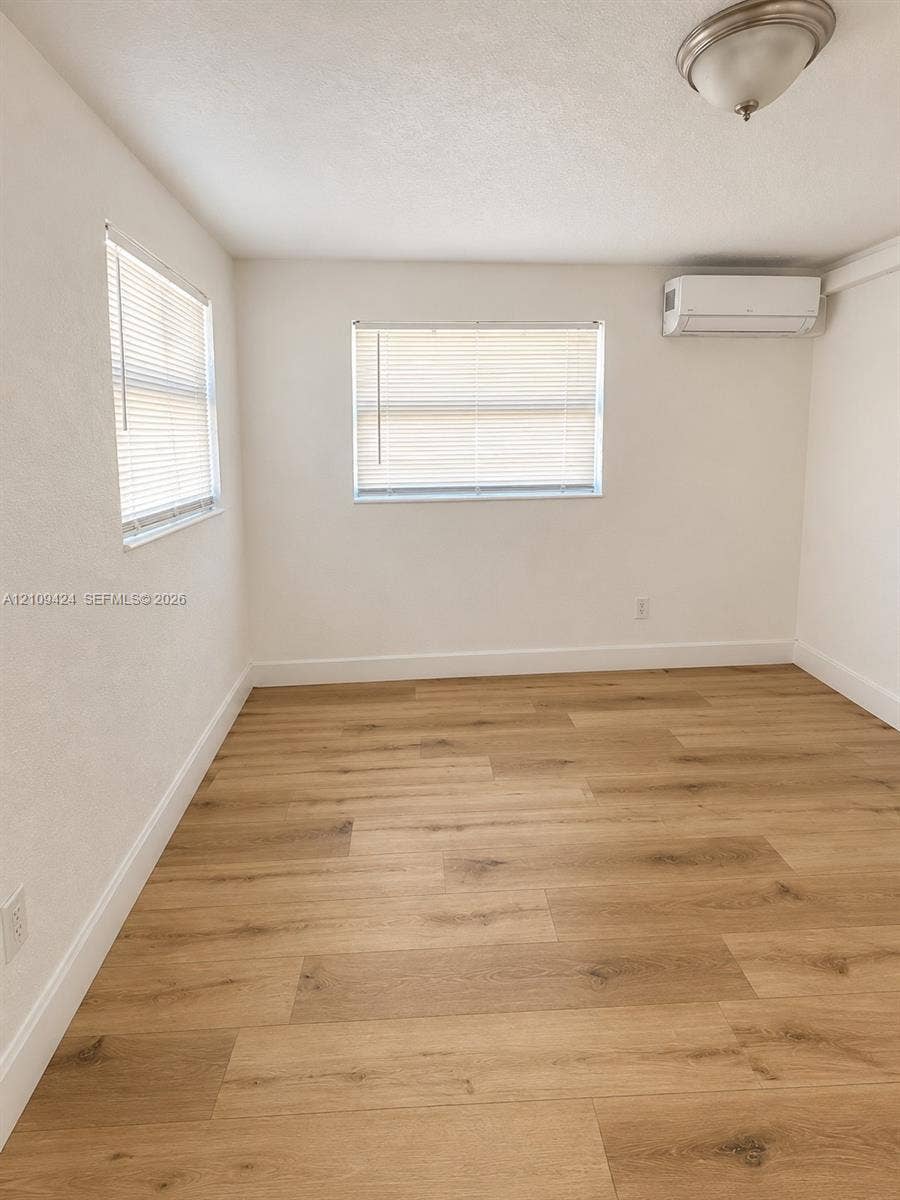 840 NW 10th Street, #5, Hallandale Beach, Hallandale Beach, FL 33009 Neutral-toned room featuring wood-finish flooring, dual windows with horizontal blinds, and a wall-mounted ductless climate control unit