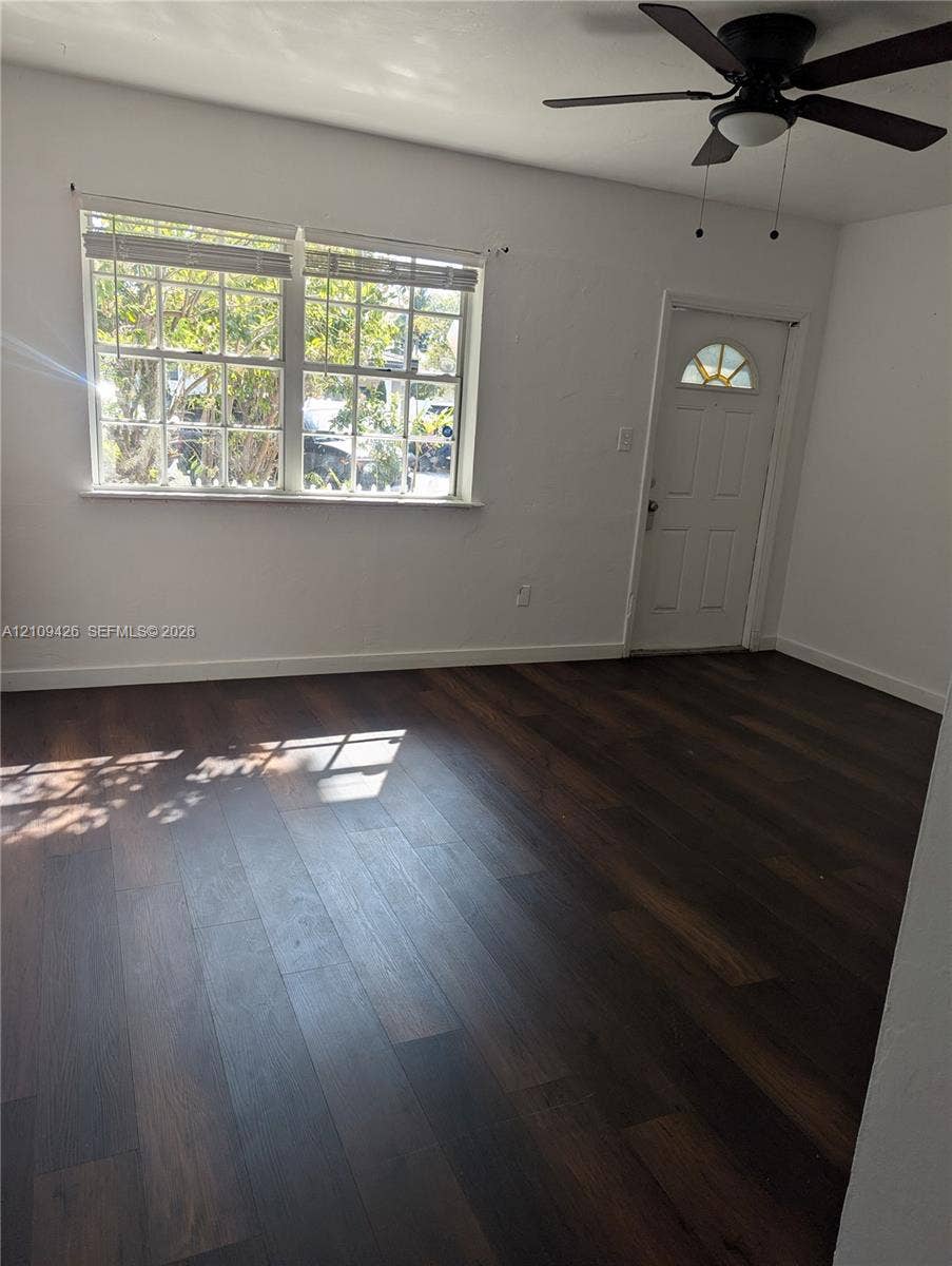 Room featuring dark wood-finish flooring, white walls, a multi-pane window, an exterior door with a semi-circular glass insert, and a ceiling fan