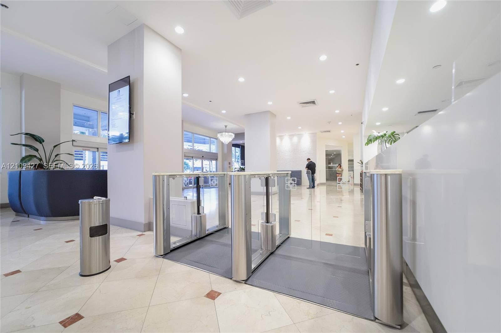 Building entrance lobby featuring polished stone flooring with decorative accents, recessed ceiling lighting, brushed metal security turnstiles, and a full-length wall mirror