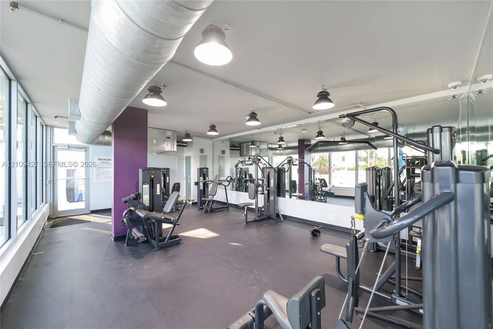 Fitness space featuring floor-to-ceiling windows, industrial exposed ductwork, wall-to-wall mirrored surfaces, and rubberized flooring