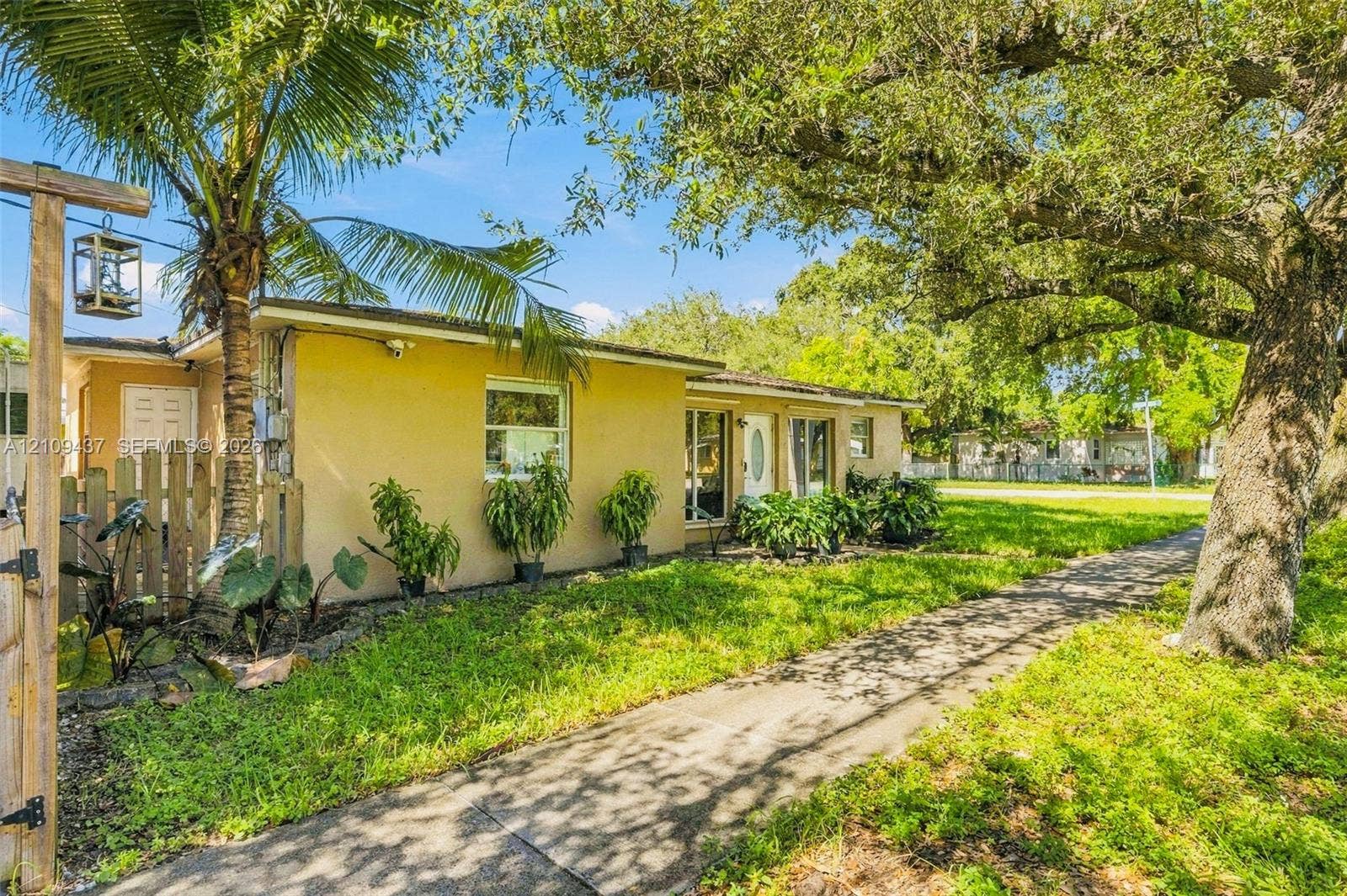 Single-story exterior featuring light-toned stucco siding, a concrete walkway, mature trees, and a wooden privacy fence