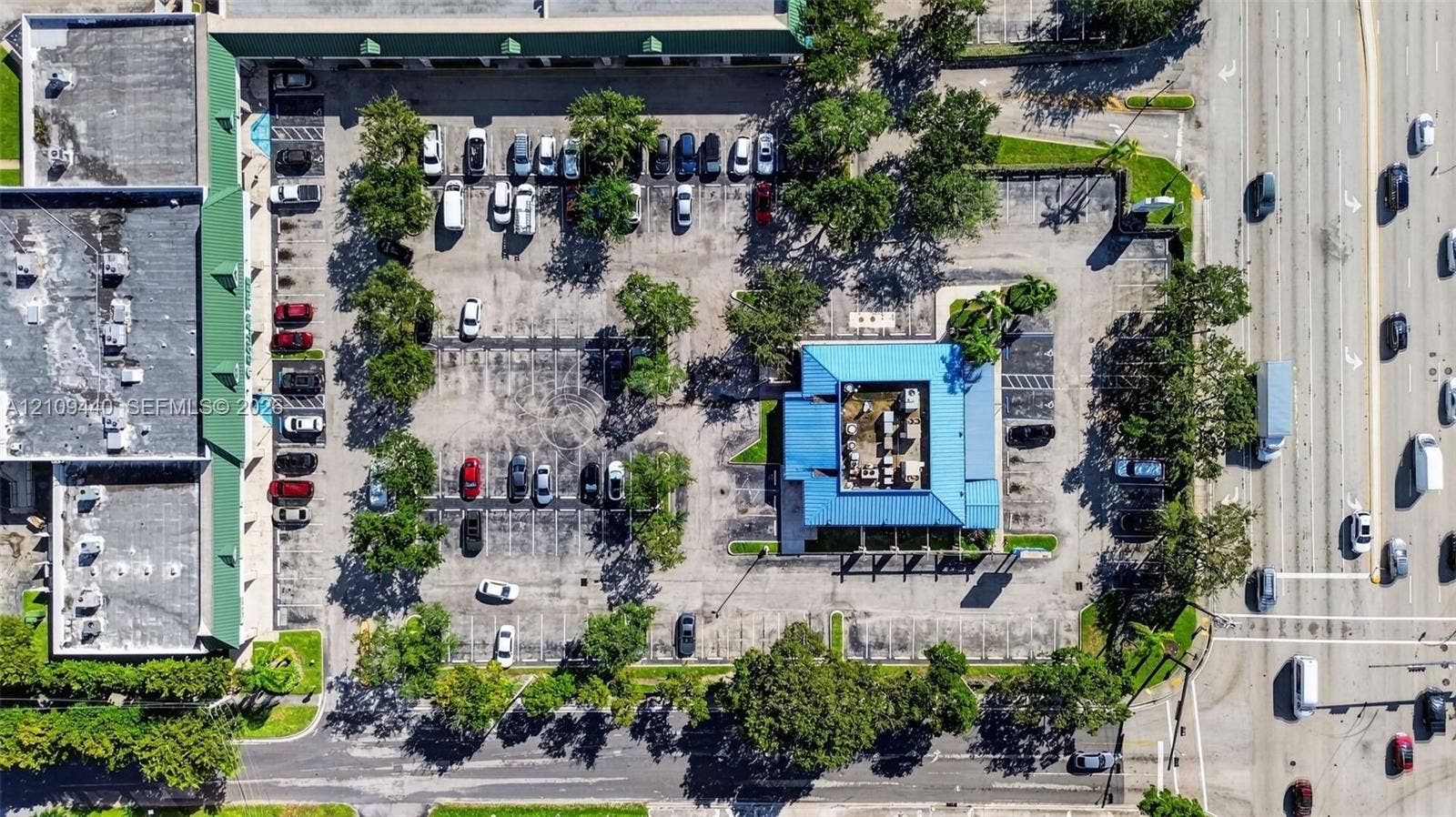 Aerial perspective featuring a commercial property with an expansive asphalt parking lot, mature shade trees, a metal roof structure, and prominent street frontage
