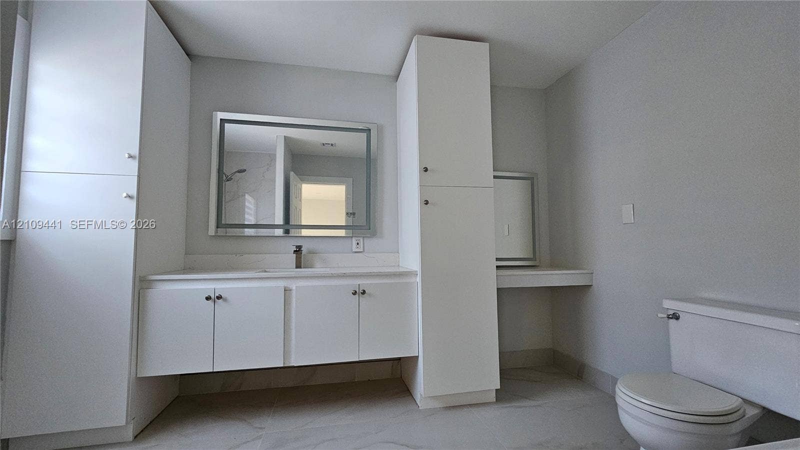 Bathroom featuring white cabinetry with integrated floor-to-ceiling storage, stone-surface countertop, rectangular wall mirror with integrated lighting, and neutral large-format tile flooring