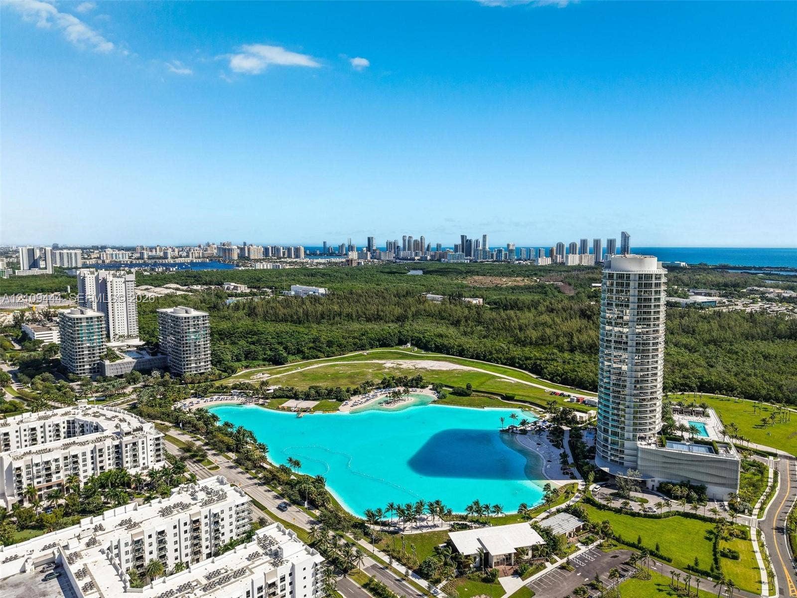 Aerial perspective of a high-rise residential tower featuring a large man-made lagoon with a sandy shoreline, surrounding parkland, and coastal city skyline