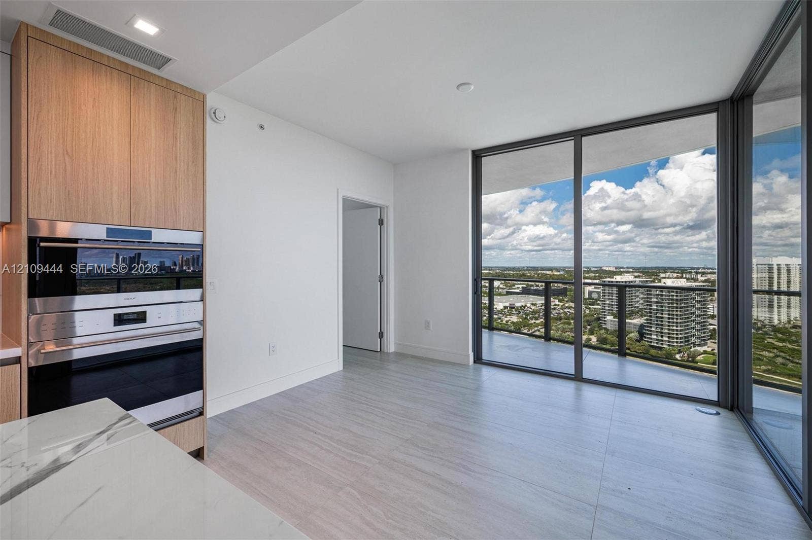Floor-to-ceiling glass sliding doors, light-toned tile flooring, built-in wall oven and microwave, custom wood-finish cabinetry, and stone-surface countertop