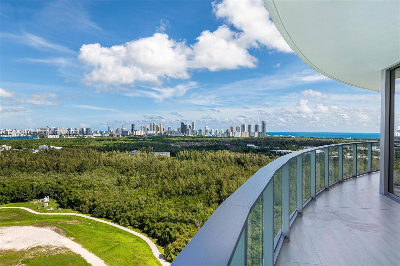 Curved exterior balcony with glass panel railing and tile flooring, featuring expansive sights of the coastal skyline and surrounding greenery