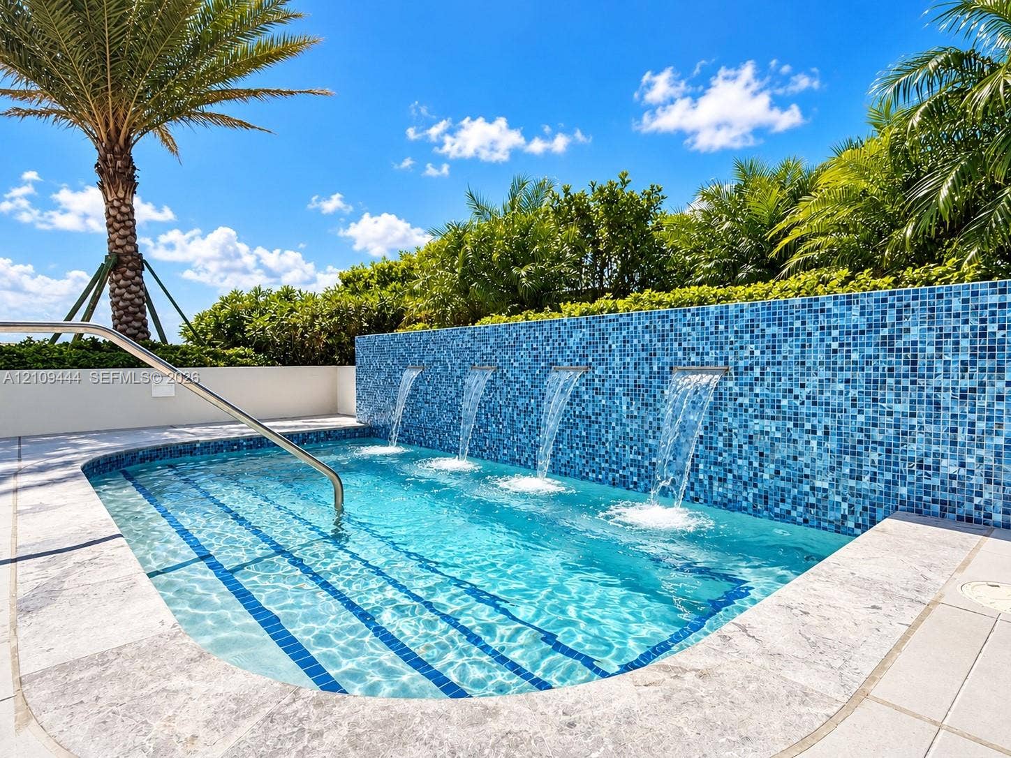In-ground pool featuring a blue glass mosaic tile water wall, four integrated water features, a metal handrail, and stone-finish coping
