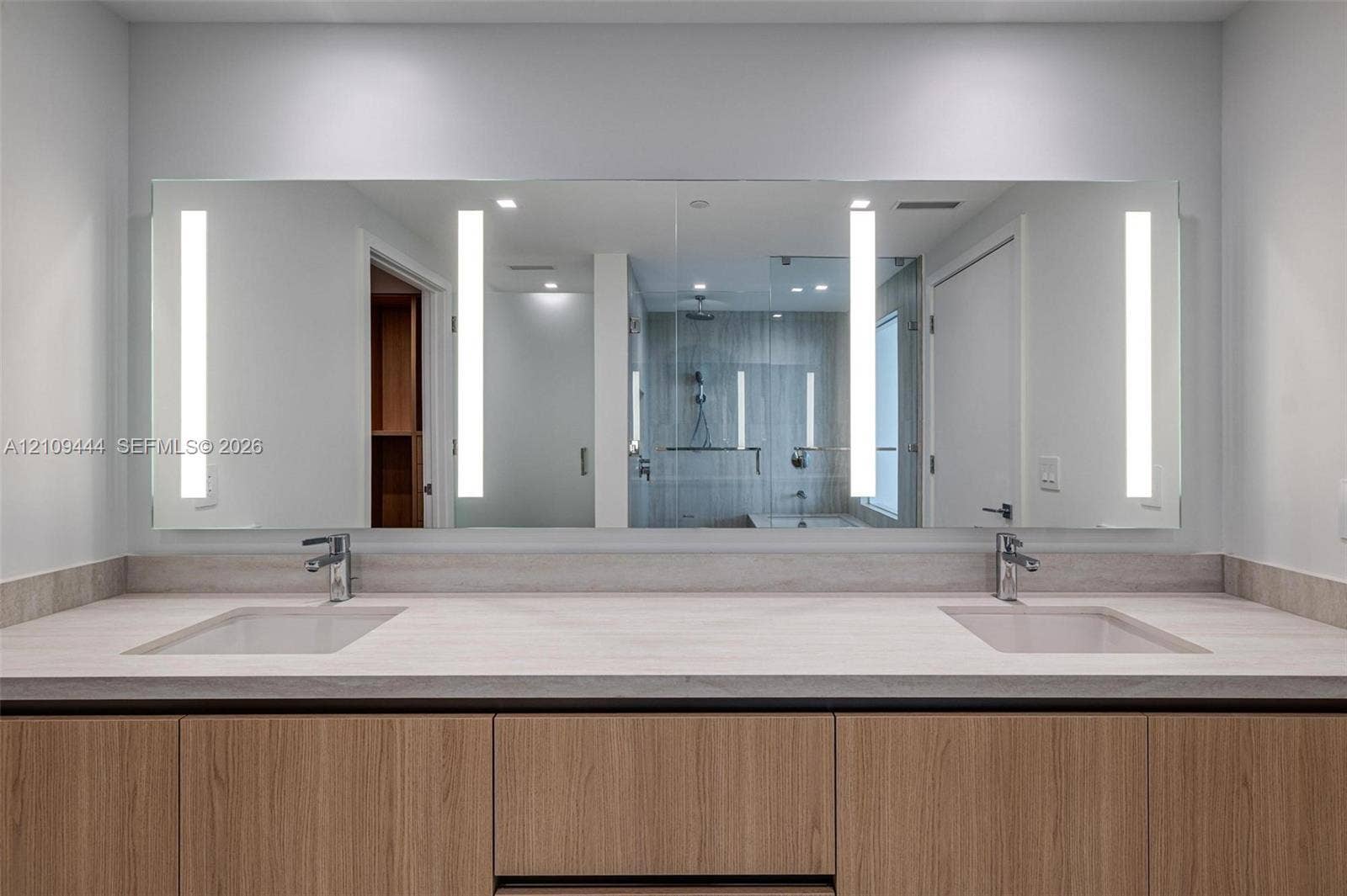 Dual-vanity cabinet with wood-finish cabinetry, neutral stone-composite countertop, integrated dual undermount sinks, and full-width mirror featuring integrated vertical LED lighting