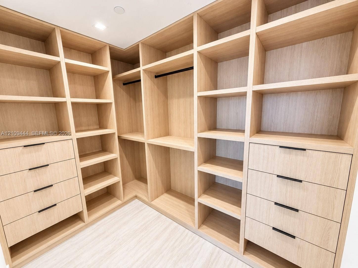 Custom walk-in storage featuring light wood-finish cabinetry, extensive built-in shelving, integrated garment rods, and two sets of drawers with matte black hardware