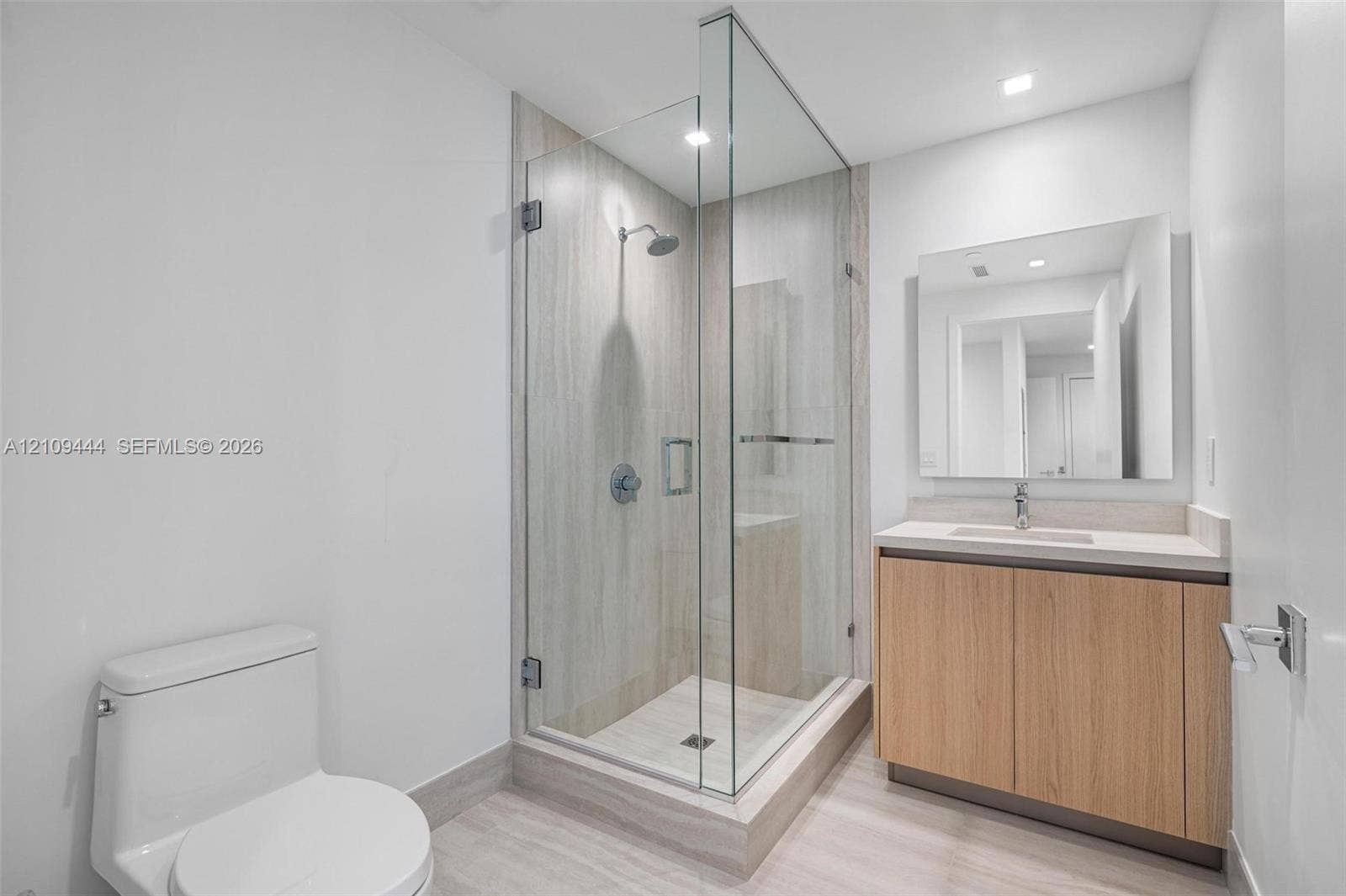 Walk-in glass shower with floor-to-ceiling tile, light wood-finish cabinetry, rectangular vessel sink, surface-mounted vanity mirror, and recessed lighting