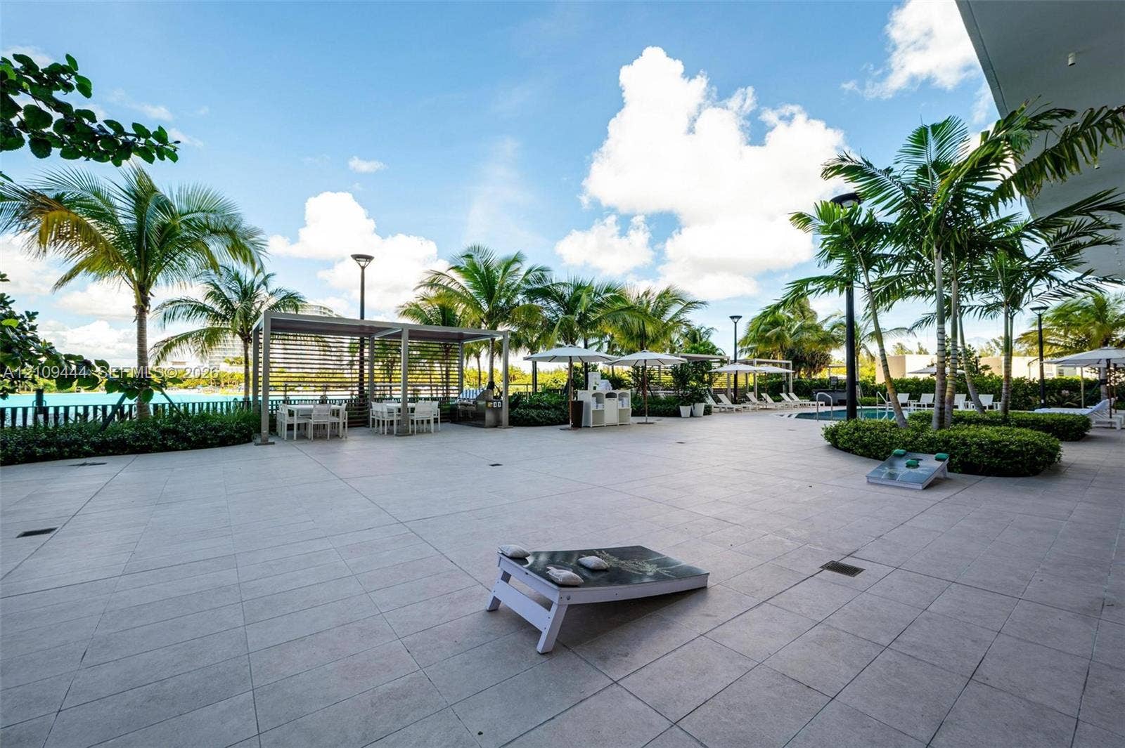 Expansive paved terrace featuring a louvered metal pergola, built-in outdoor kitchen, integrated landscape lighting, and mature palm trees