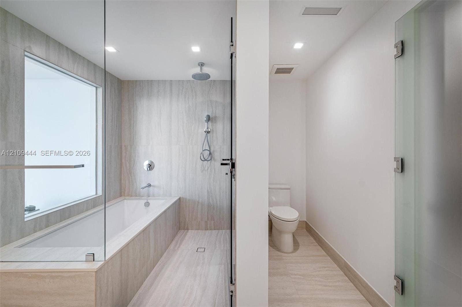 Bathroom featuring floor-to-ceiling stone-finish tile, glass-enclosed soaking tub with rain-style shower head, recessed lighting, and a private water closet