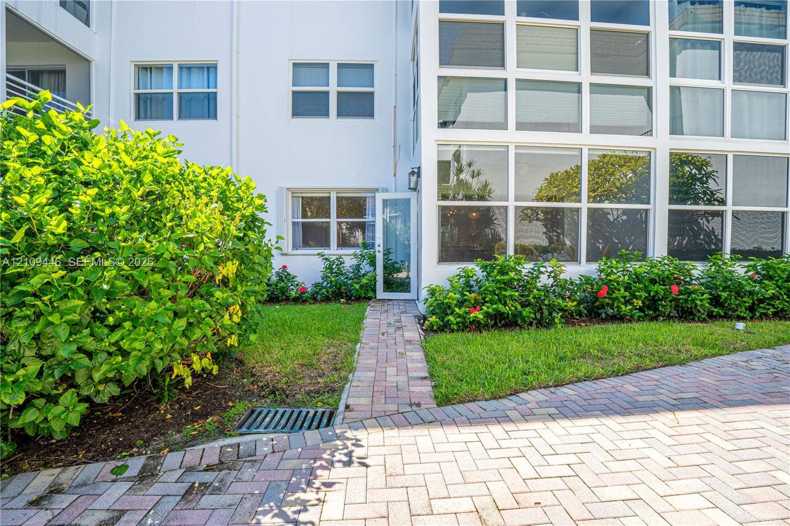 1501 S Ocean Boulevard, #117, Lauderdale-By-The-Sea, Lauderdale By The Sea, FL 33062 Exterior facade featuring white siding, a multi-pane glass wall, brick-style paver walkway, and manicured landscaping